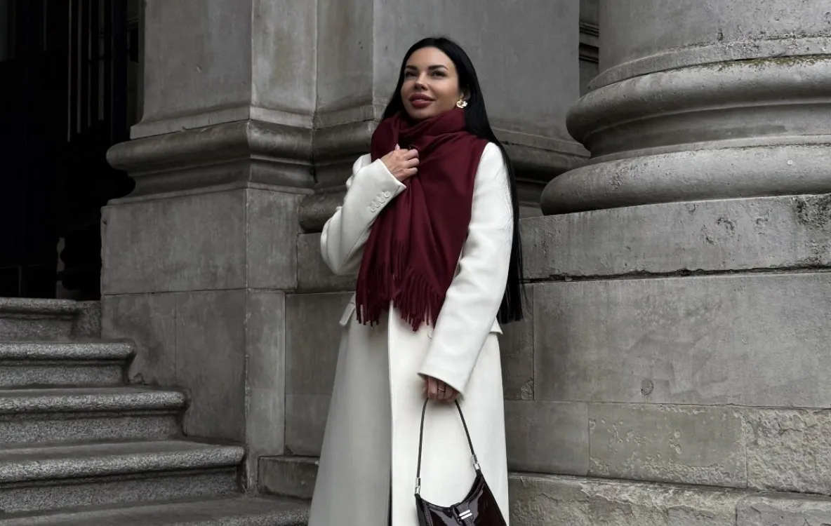 Iryna Ribeiro in a white coat with a burgundy scarf by the columns of a building