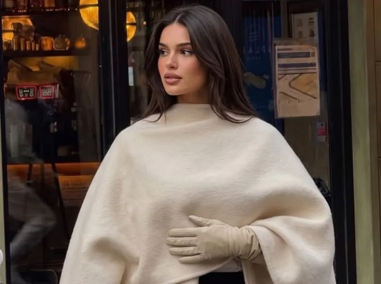 A woman in a cream poncho and gloves on the street outside a café