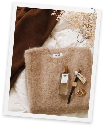 A knitted jumper, perfume and gold jewellery laid out on a bed